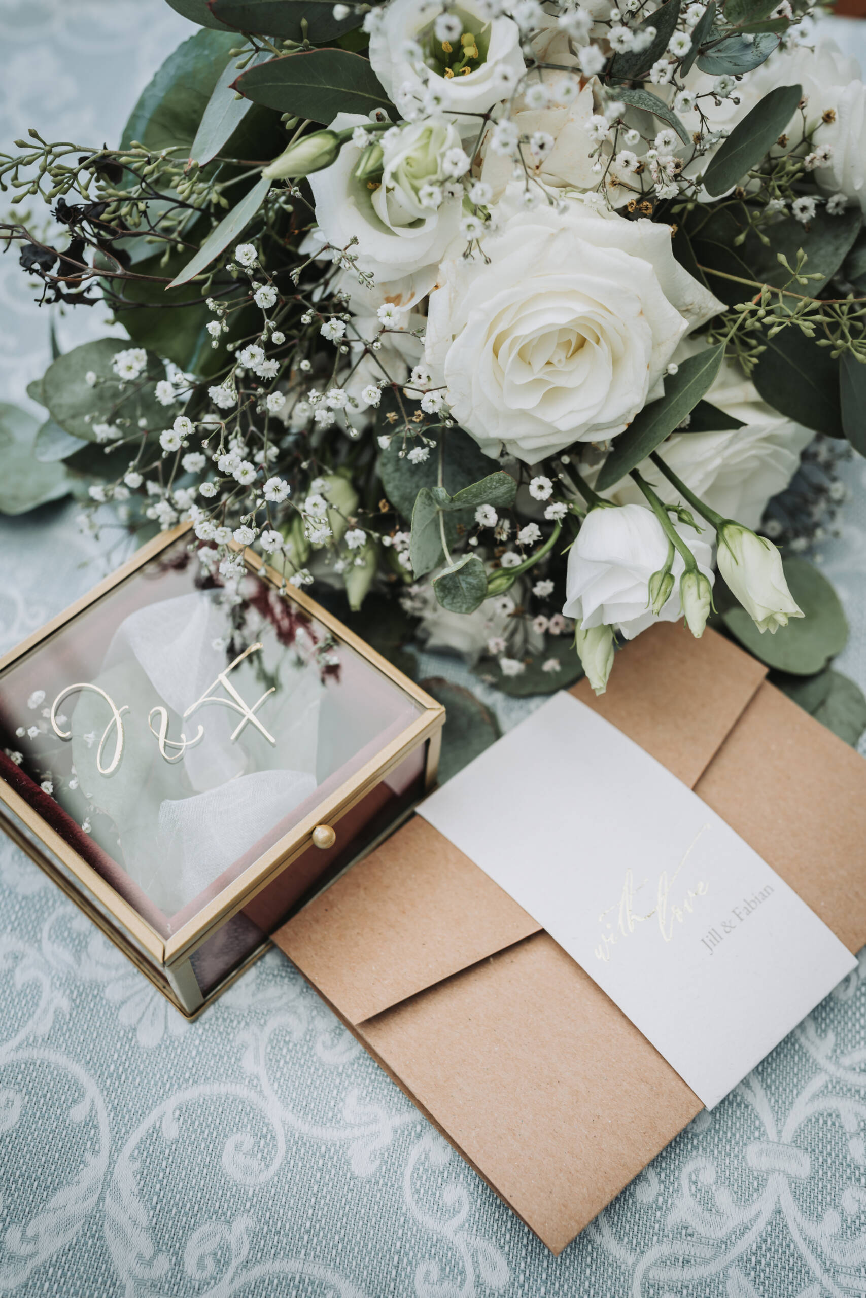 Eine Detailaufnahme einer Schmuckschatulle in welchem sich die Eheringe des Brautpaares befinden. Im Hintergrund befindet sich der wunderschöne weiß grüne Brautstrauß der Braut.