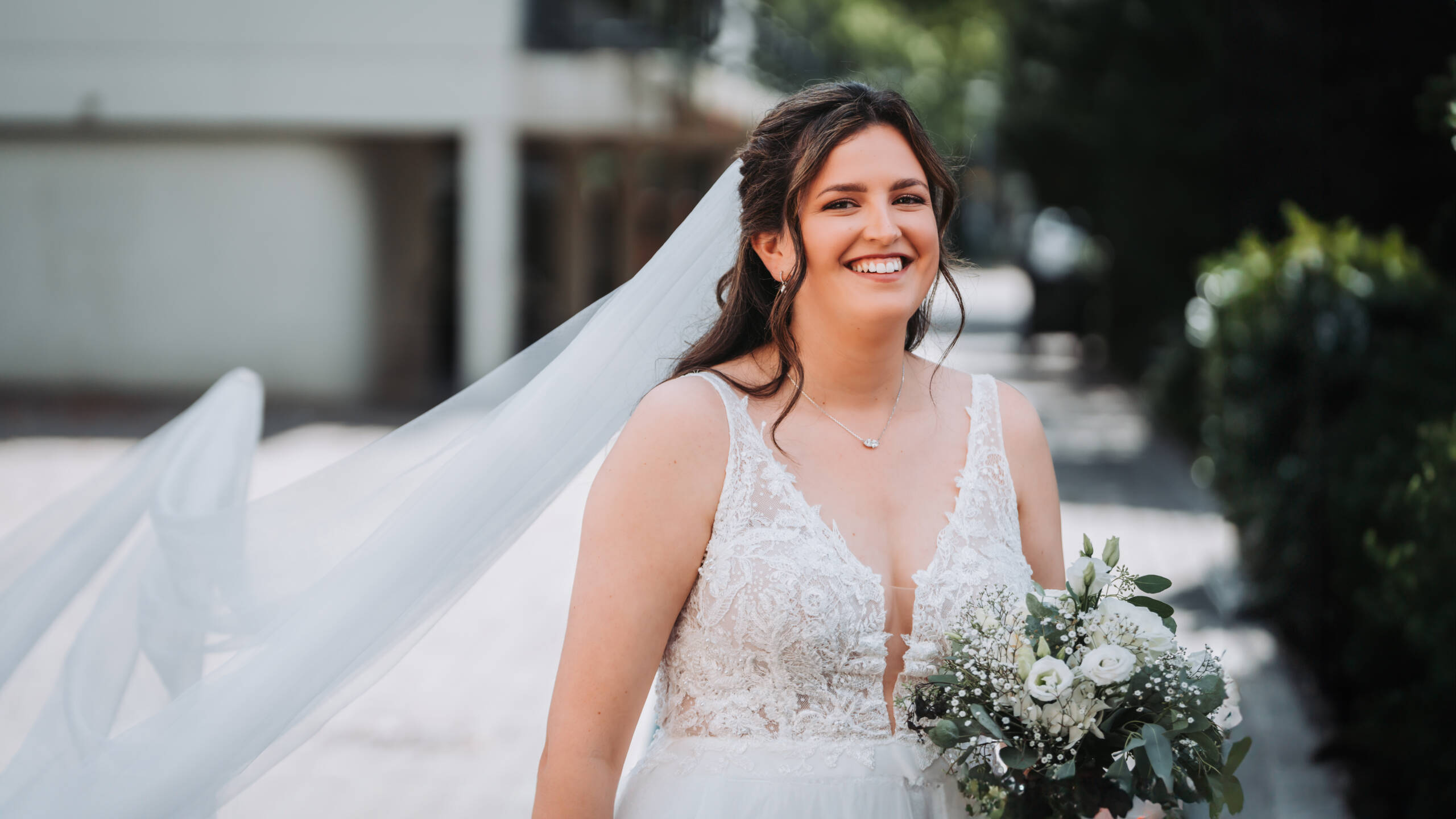 EIne Braut lächelt in die Kamera und steht kurz vor ihrer Kirchlichen Trauung. Der weiße Schweif ihres Kleides weht im Wind und die Braut hält ihren Brautstrauß in der Hand.