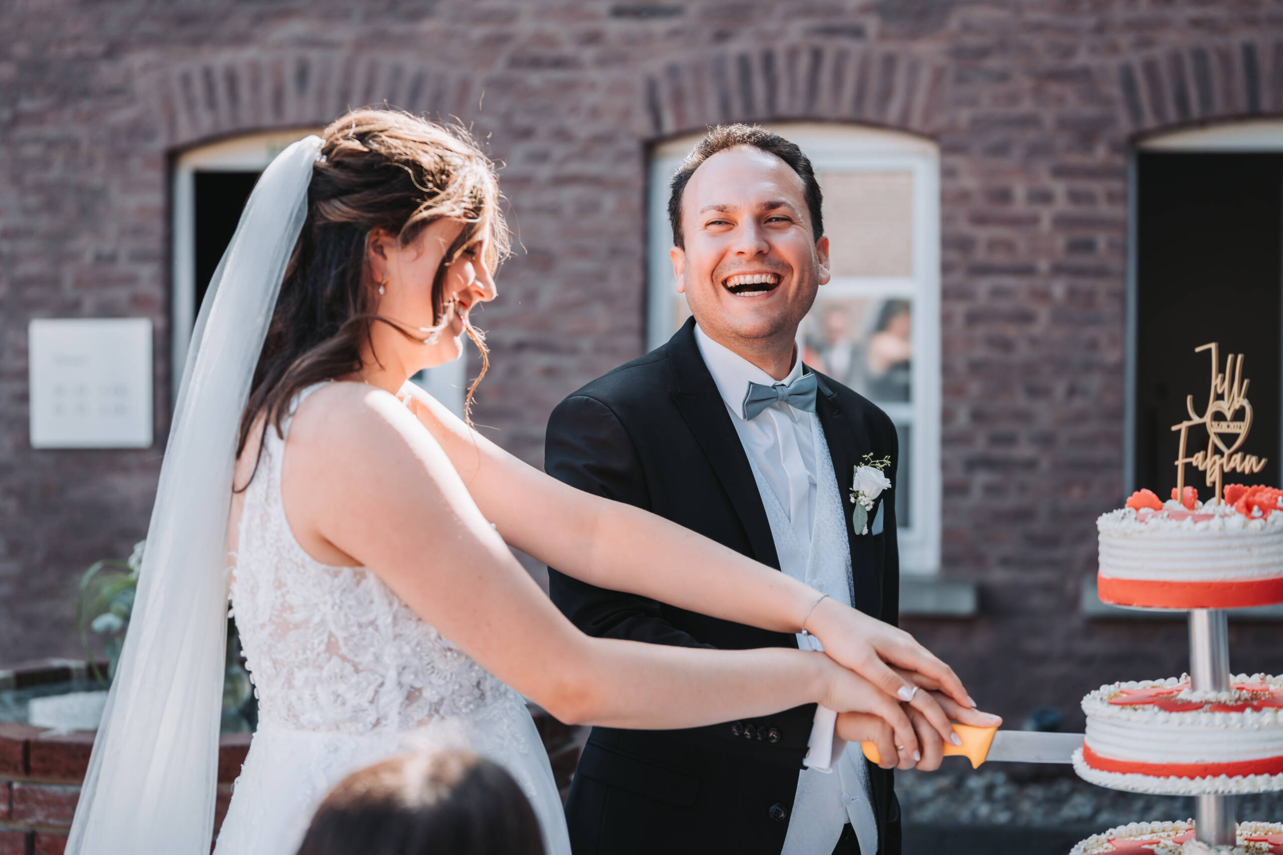 Eine Nahaufnahme von Jill und Fabian, welche gemeinsam ihre Hochzeitstorte mit einem langen Messer Anschneiden und dabei sichtlich spaß haben und lachen.