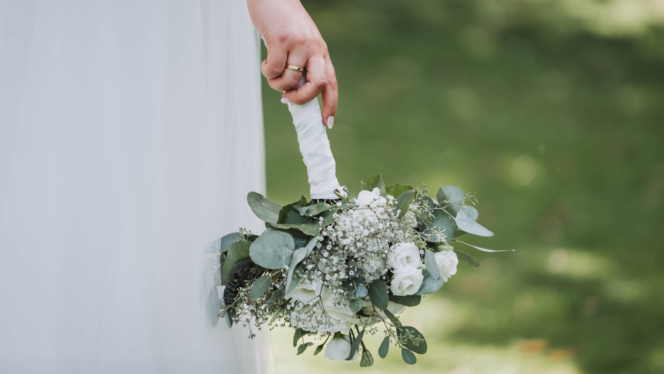 eine Braut hält ihren Brautstrauß neben ihr Hochzeitskleid nach Unten. Im Hintergrund erkennt man eine grüne Wiese.