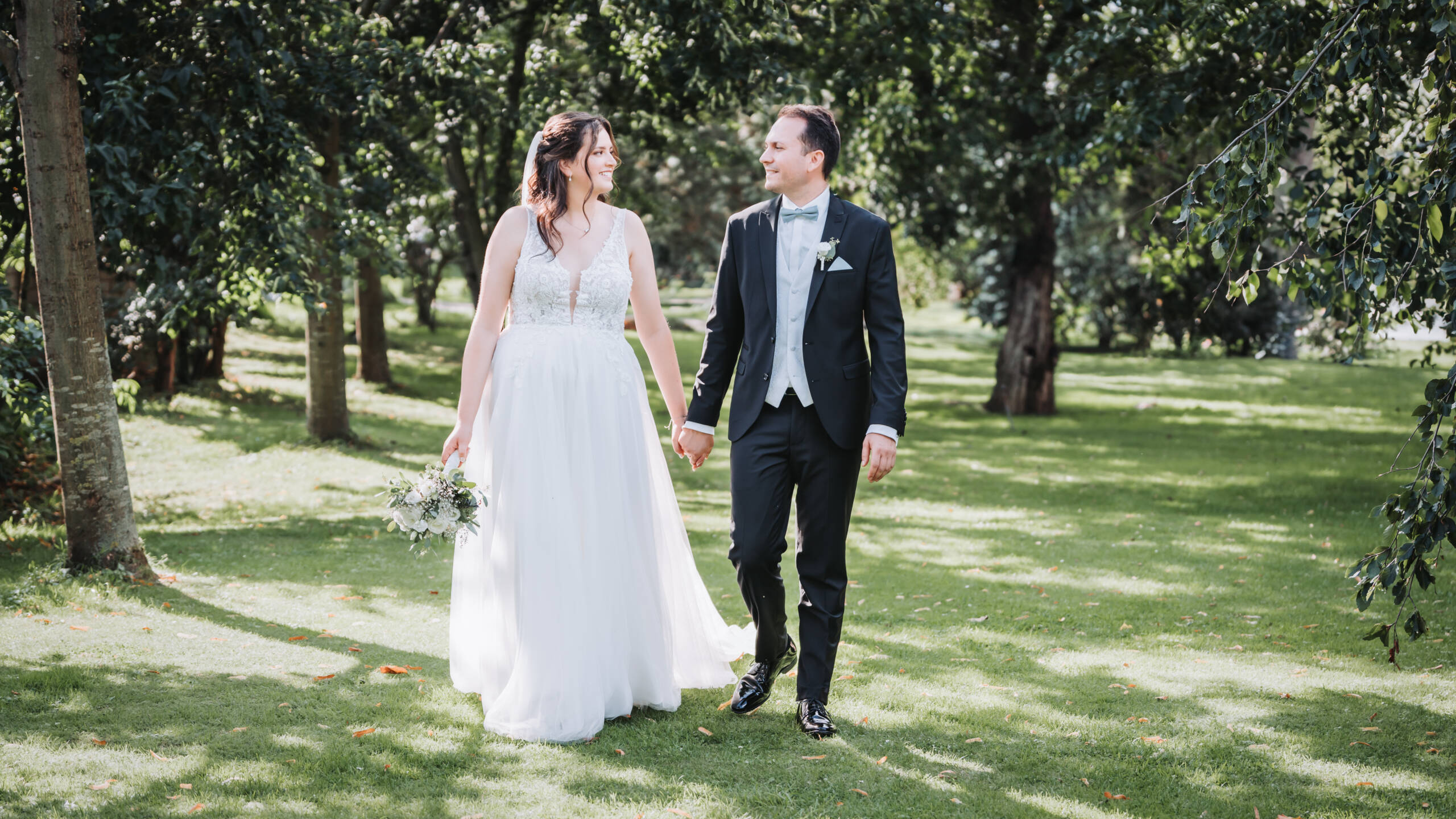 Ein frisch verheiratetes Hochzeitspaar läuft auf einer grünen Wiese durch eine Allee von Bäumen und genießt das beste Wetter und lässt Hochzeitsfotos von einem Hochzeitsfotografen auf Neuss von sich anfertigen.
