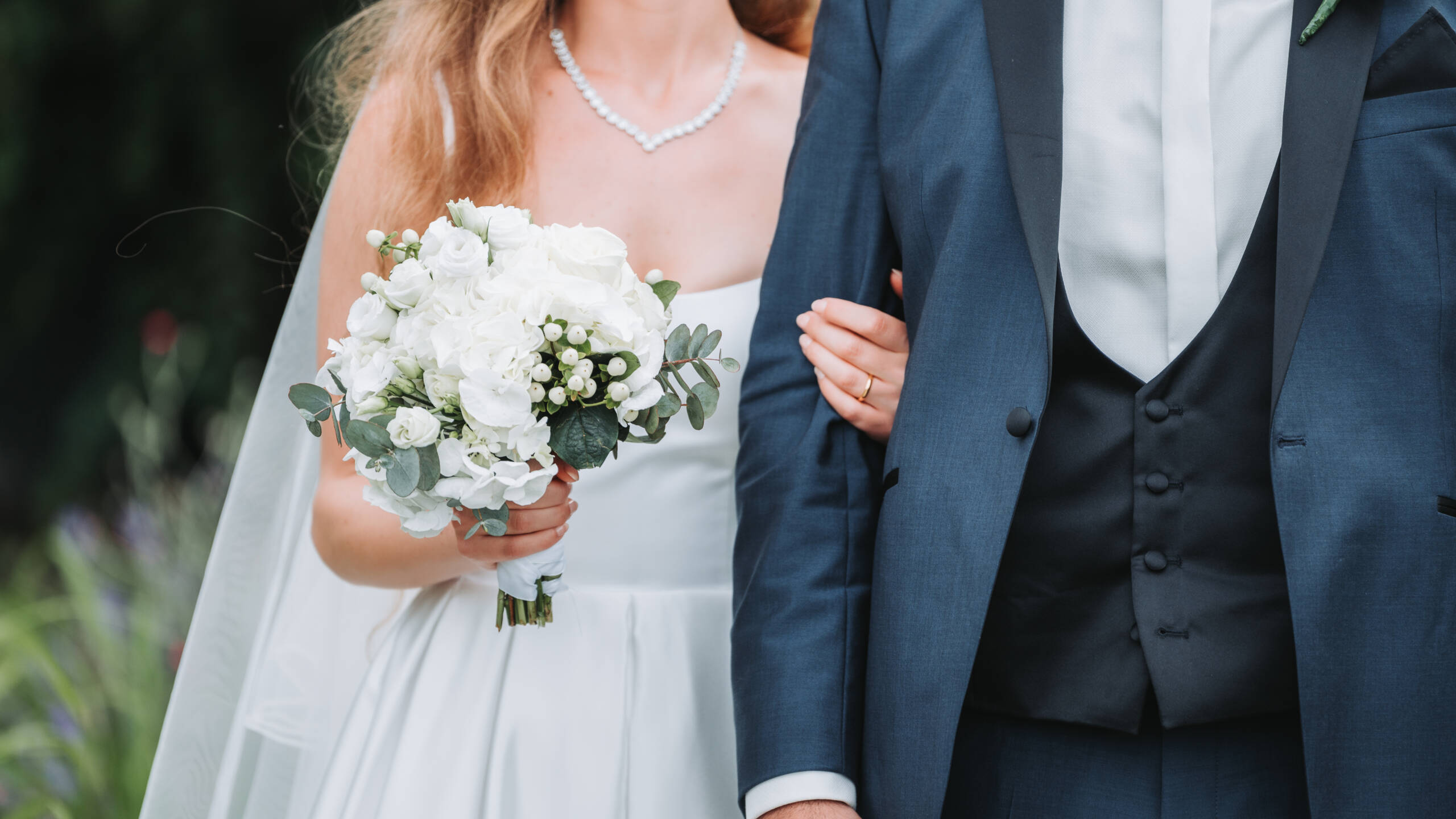 Eine Braut hält sich am Arm ihres Bräutigams fest und trägt einen Brautstrauß in der anderen Hand. Das Foto ist während des Brautpaarshootings kurz nach der kirchlichen Trauung entstanden.