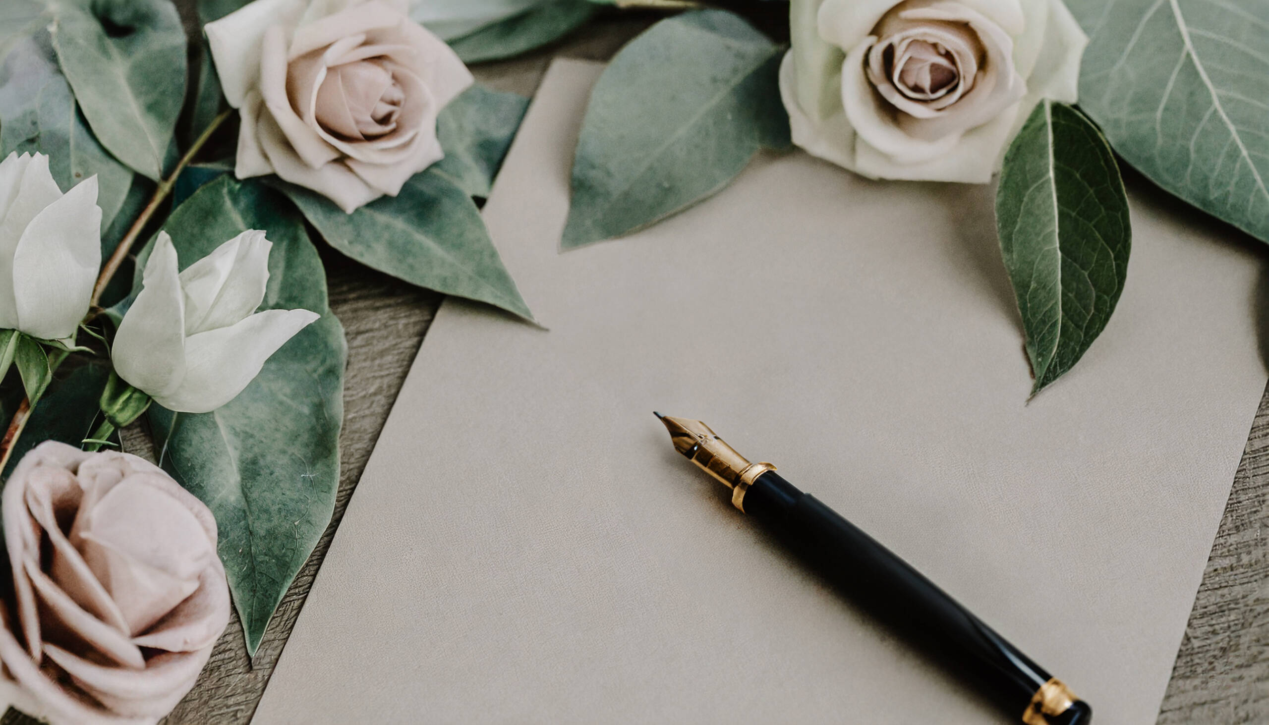 Ein Füllfederhalter liegt auf einem hochwertigen Papier umgeben von weißen und beigen Rosen auf grünen Blättern. Das ist Teil einer Hochzeitsdecoration für die Webseite eines Hochzeitsfotografen aus Neuss.